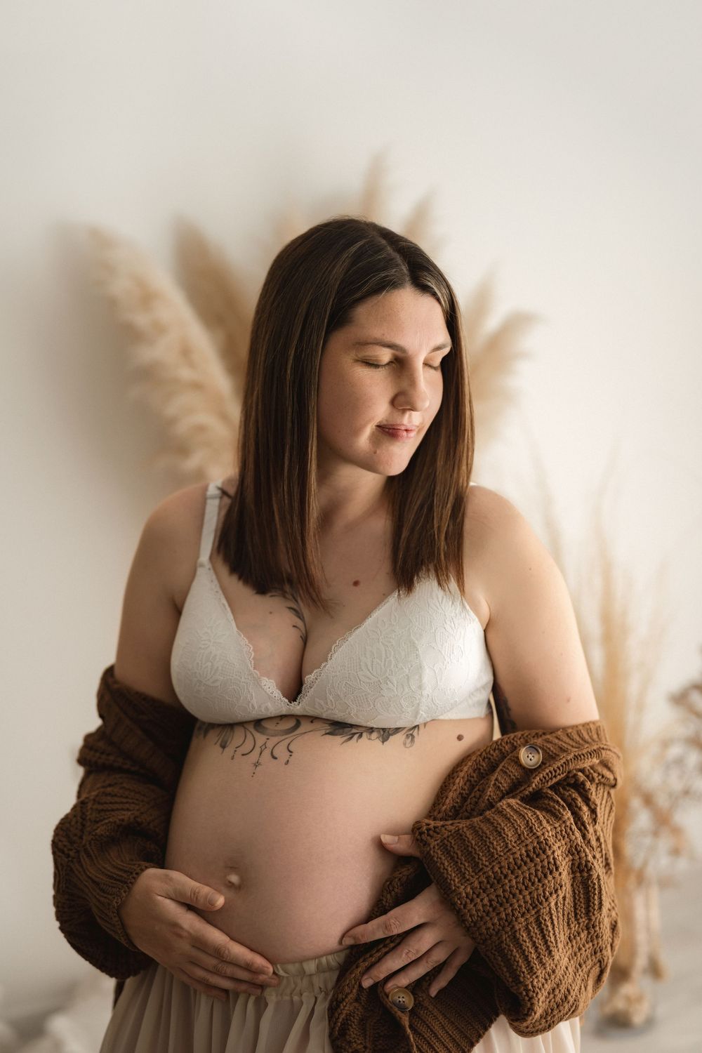 Pregnant woman in white lace bra and brown cardigan cradling baby bump, with pampas grass decor in background.