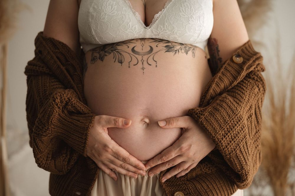 Pregnant woman cradling baby bump, floral tattoo, white bra, brown knit cardigan, maternity photo.