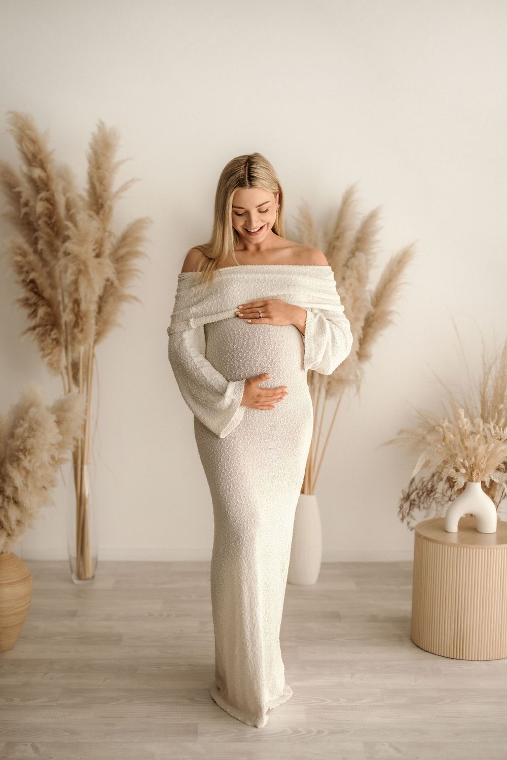 Pregnant woman in white off-shoulder knit dress cradling belly, surrounded by pampas grass decor in a neutral studio.