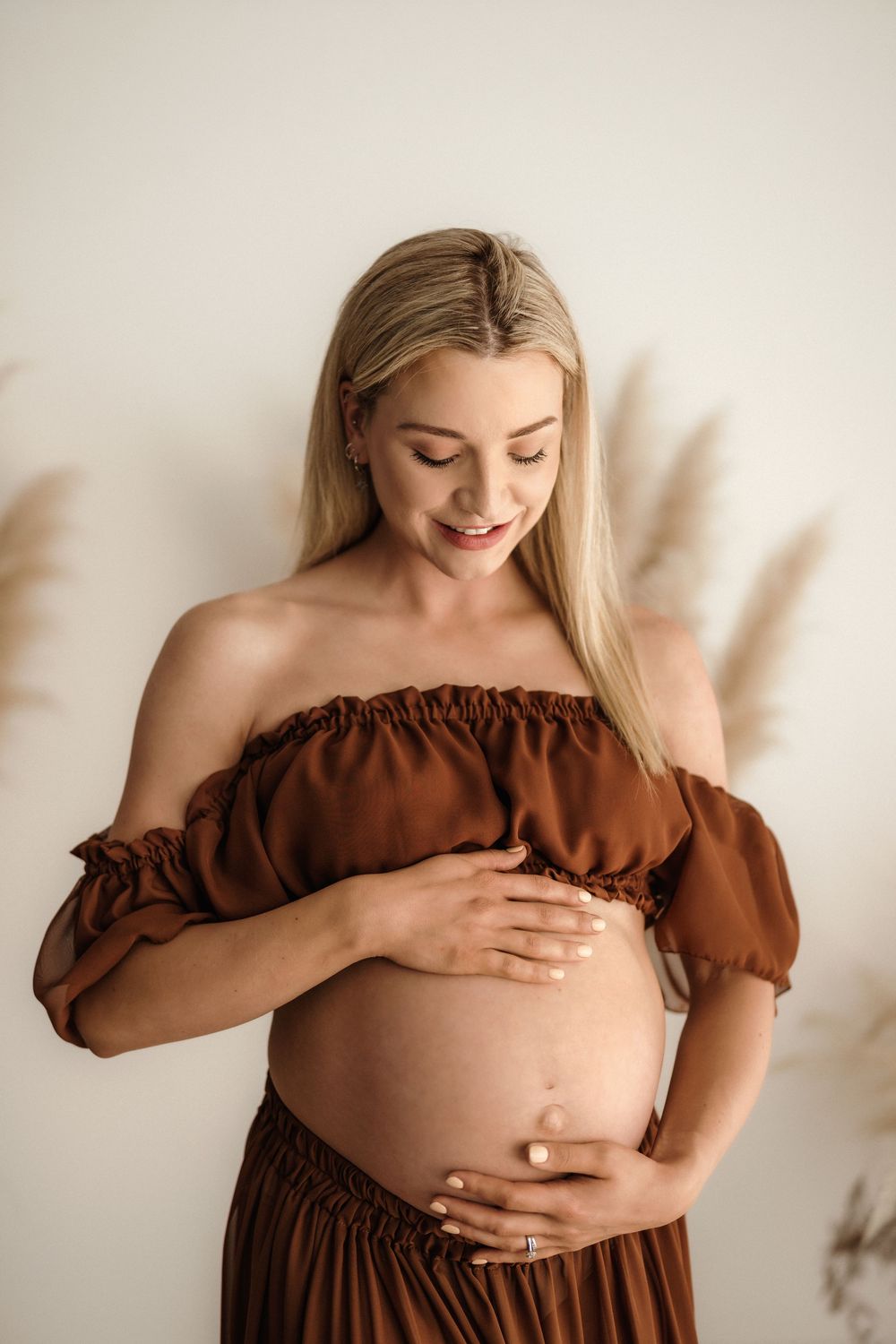 Smiling pregnant woman in brown off-shoulder dress cradling her baby bump against a neutral background.