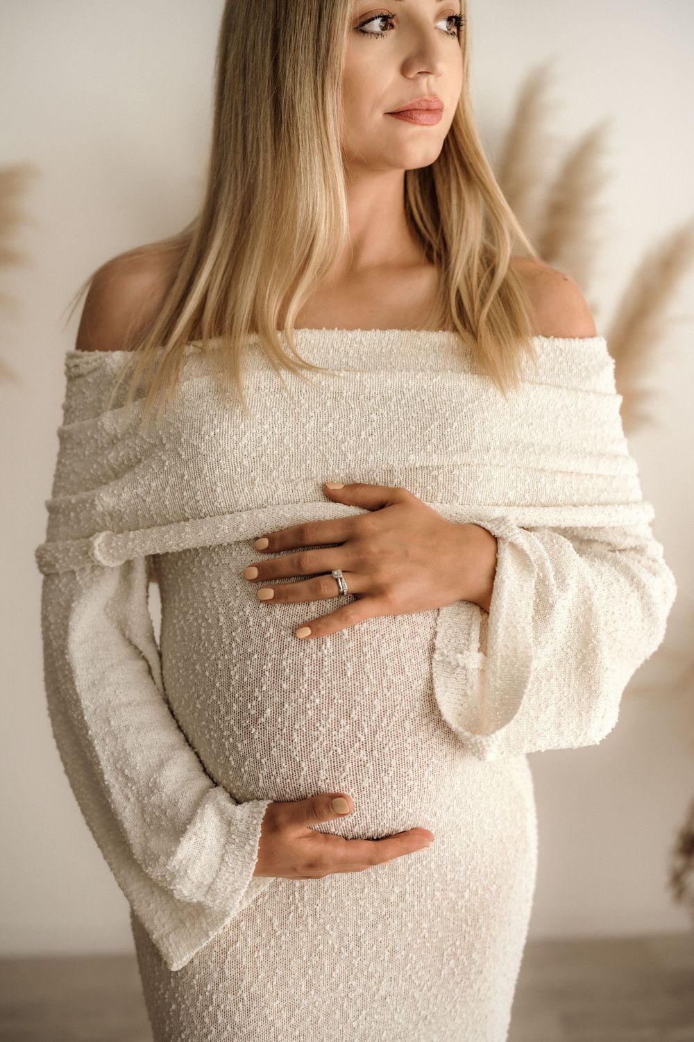 Pregnant woman in white off-shoulder dress cradling baby bump, wearing engagement ring, soft neutral background.