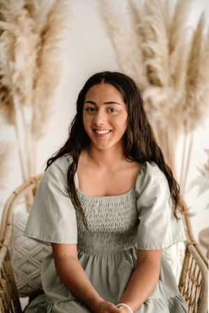 Portrait of someone wearing a sage green smocked dress against a wall decorated with pampas grass.