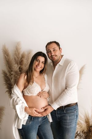 Casual maternity photo with couple in white tops and jeans against neutral background with pampas grass.