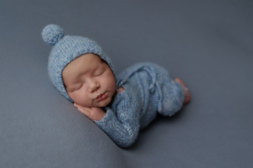 A newborn boy in a cozy blue knitted outfit, peacefully resting on his side.