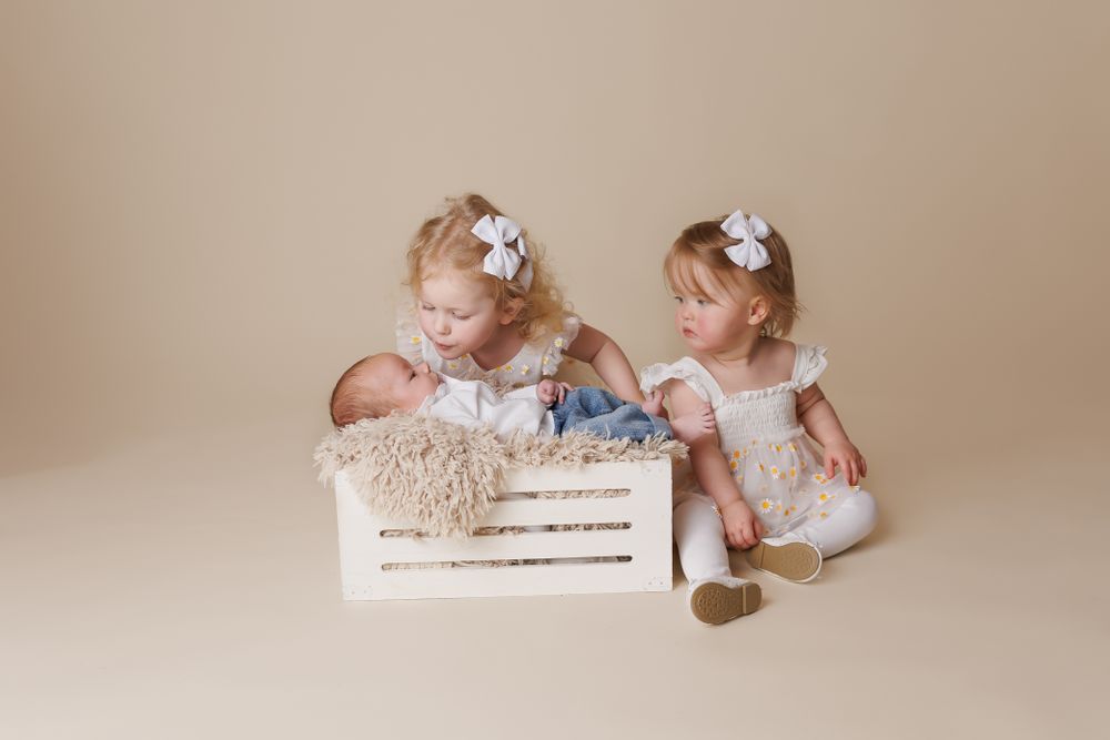 two older sisters looking lovingly at their newborn baby sister on a cream background