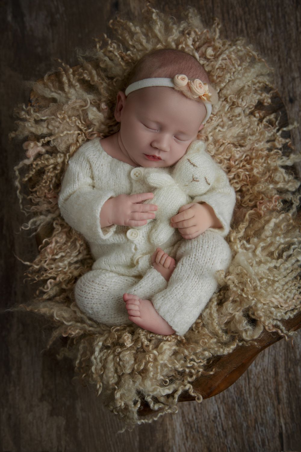 A peaceful newborn girl in a white outfit peacefully sleeps on a cozy wooden chair.