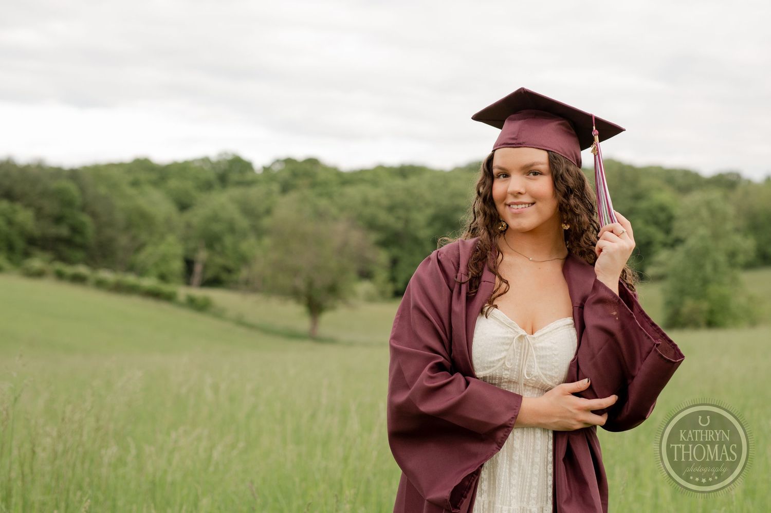 Graduate in maroon regalia and white dress poses confidently in green meadow.