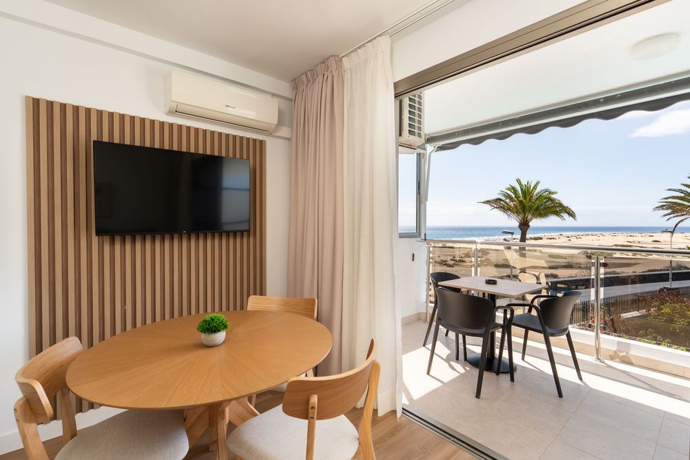 Modern coastal apartment interior with round wooden table, chairs, and panoramic ocean views through large windows.