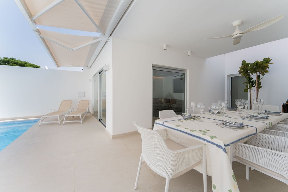 Bright white outdoor dining area with long table and chairs beside a sparkling pool and covered patio.