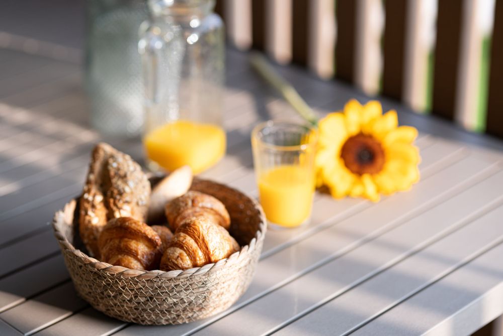 Breakfast spread with croissants in woven basket, orange juice and sunflower on wooden table.