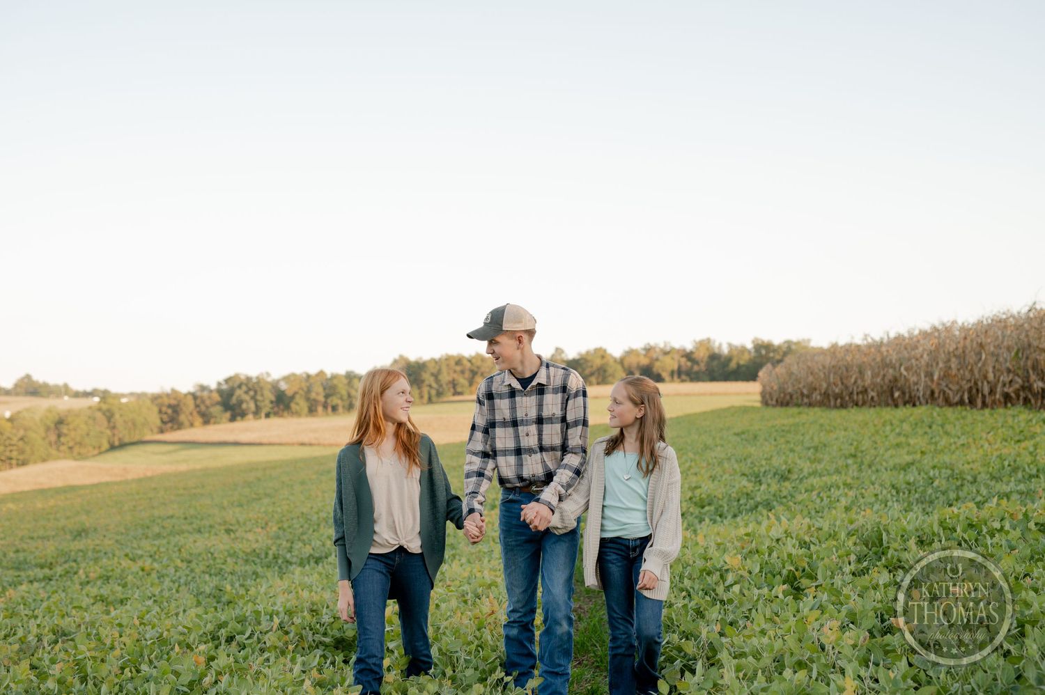 Three people stand together in a sunny farm field with cornfields in the background.