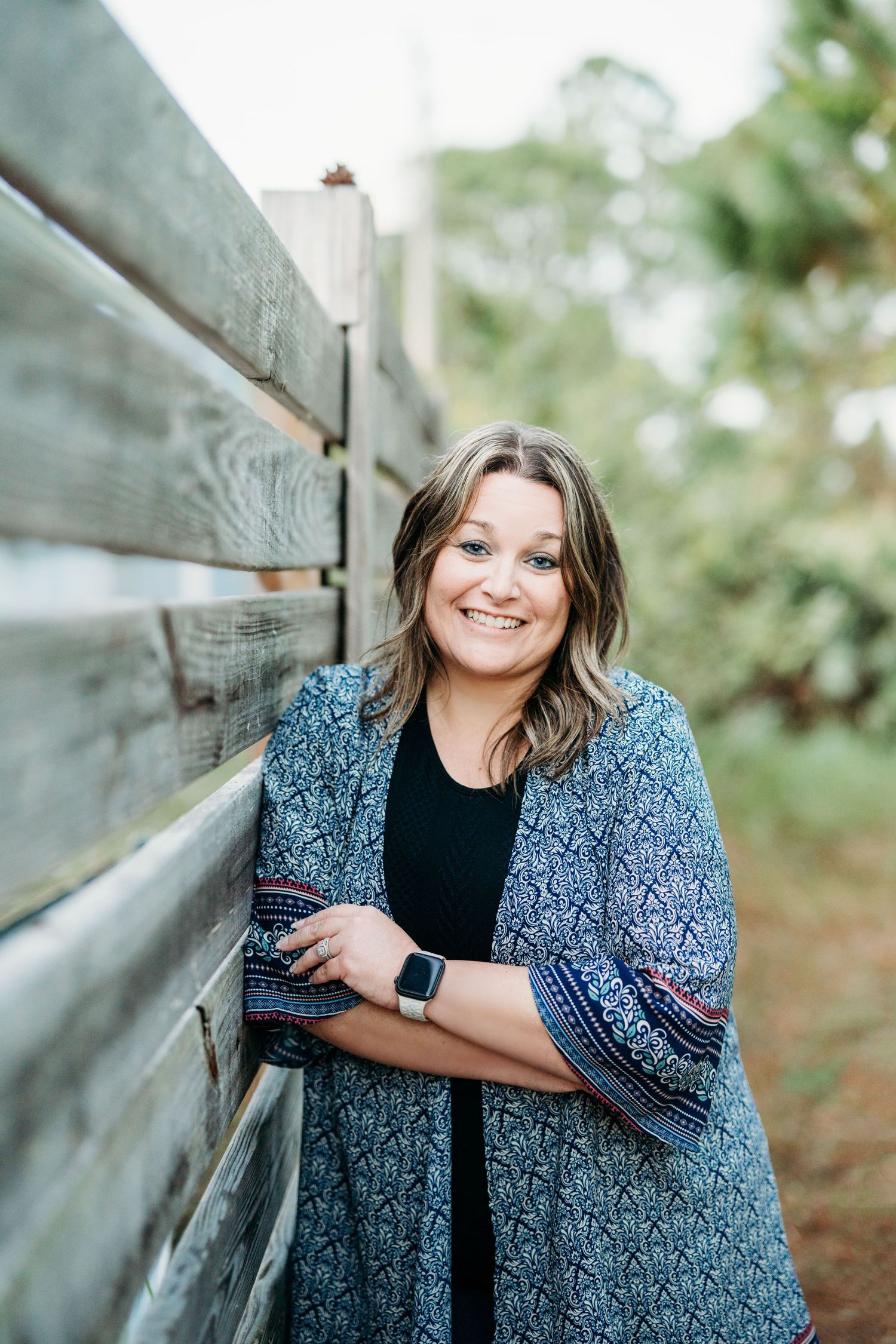 A woman in a patterned cardigan leans against a wooden fence while wearing a smartwatch and smiling outdoors.