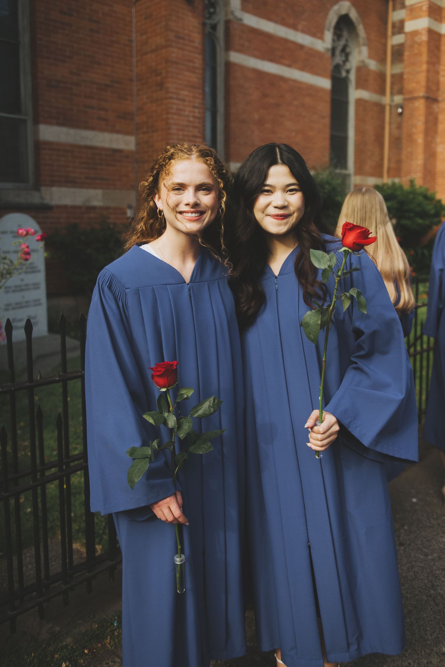 Two graduates in blue commencement robes hold red roses while standing outside a brick building.