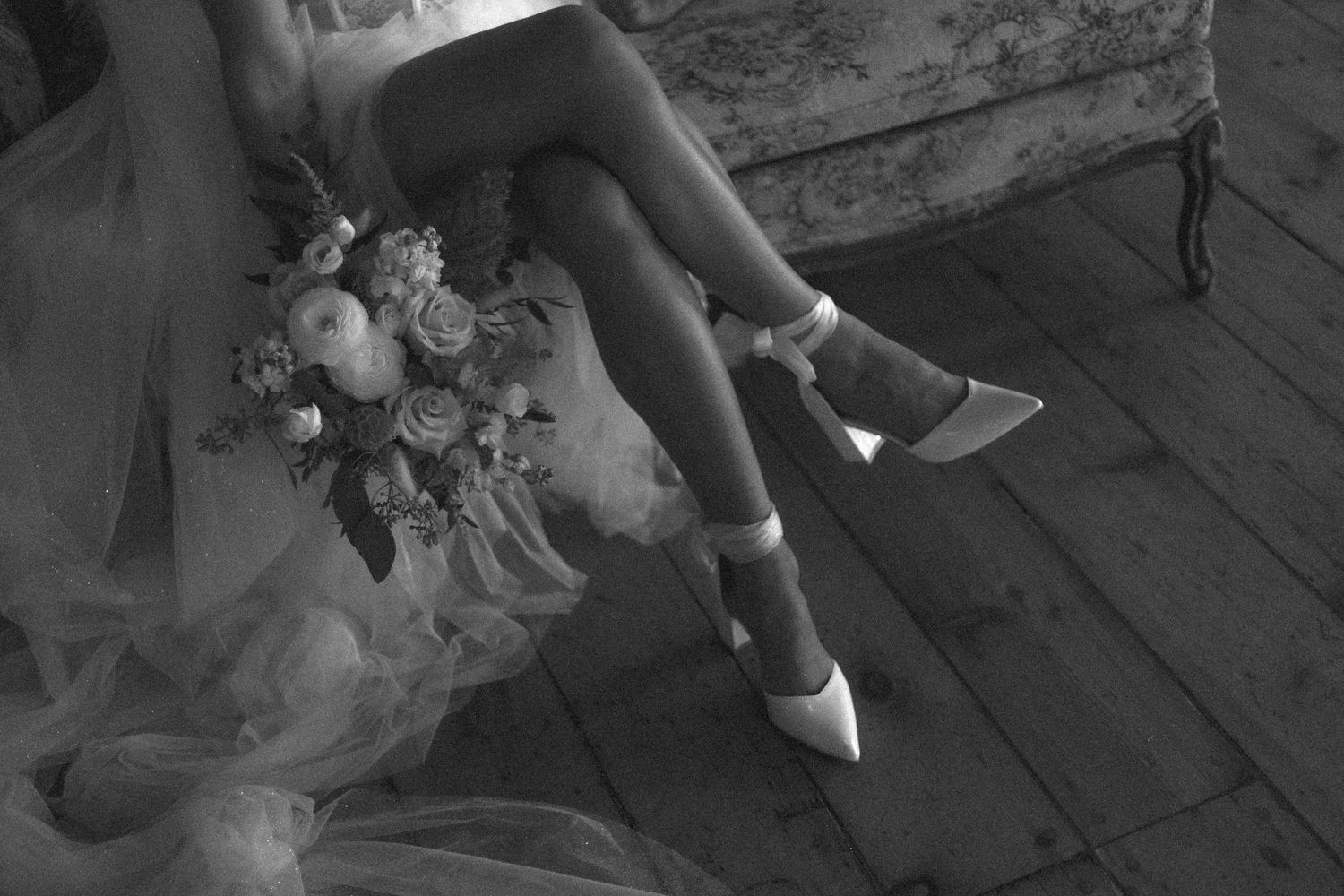 A black and white detail shot of white heels and flowing dress fabric against wooden floorboards.