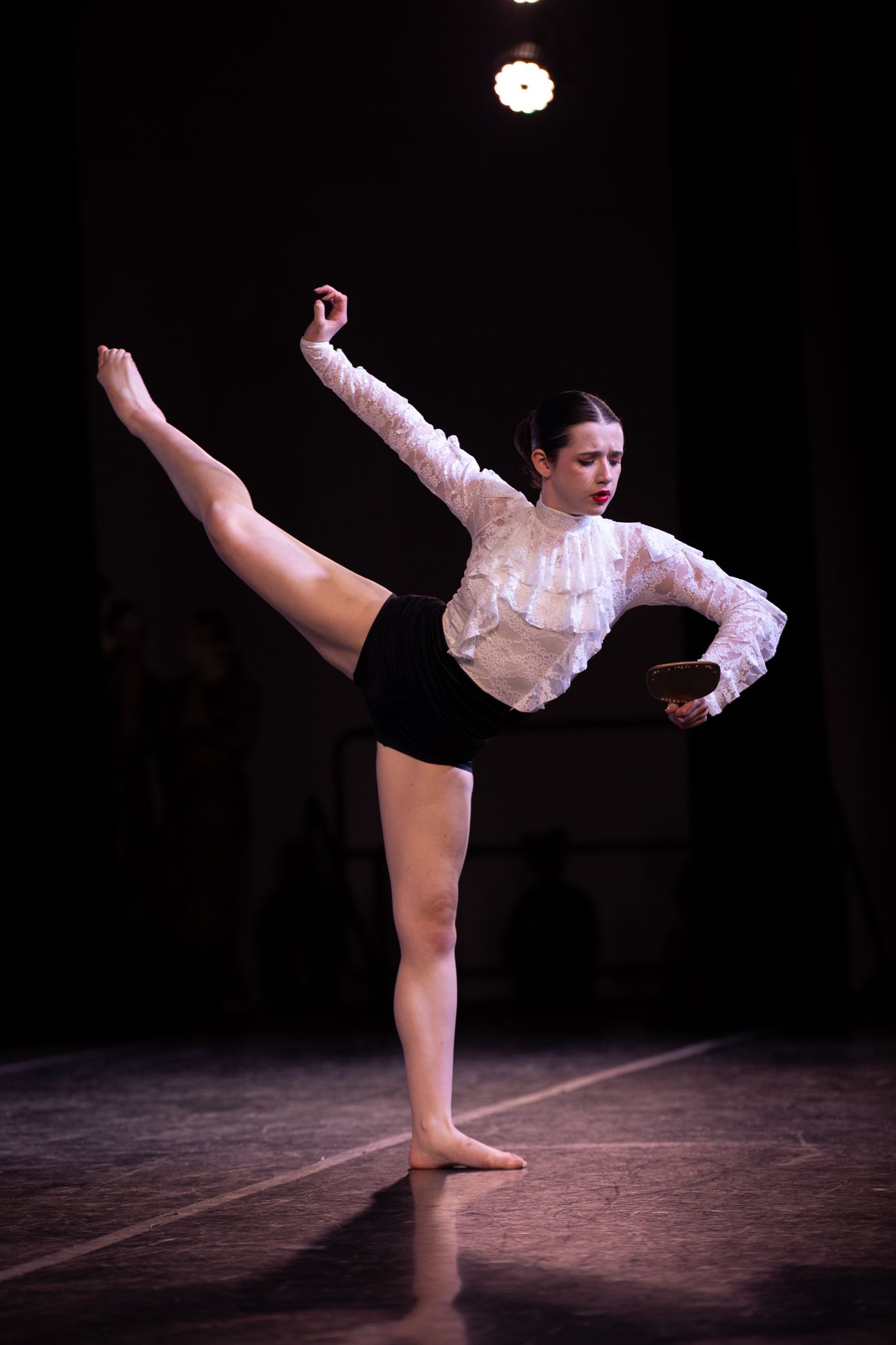 Dancer in white lace top and black shorts performs high leg extension on dark stage.