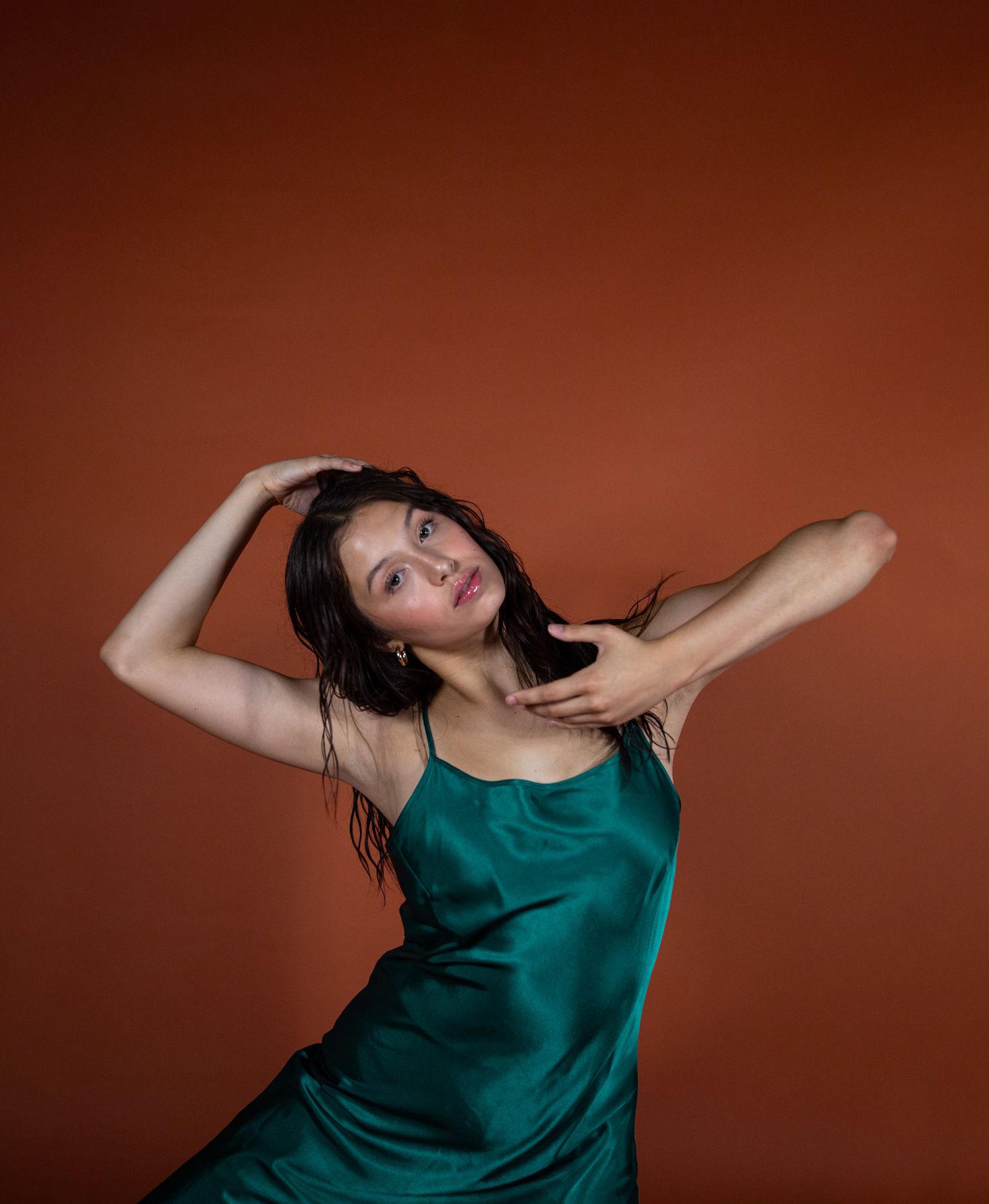 Dancer in emerald silk dress poses expressively against rust-colored backdrop.