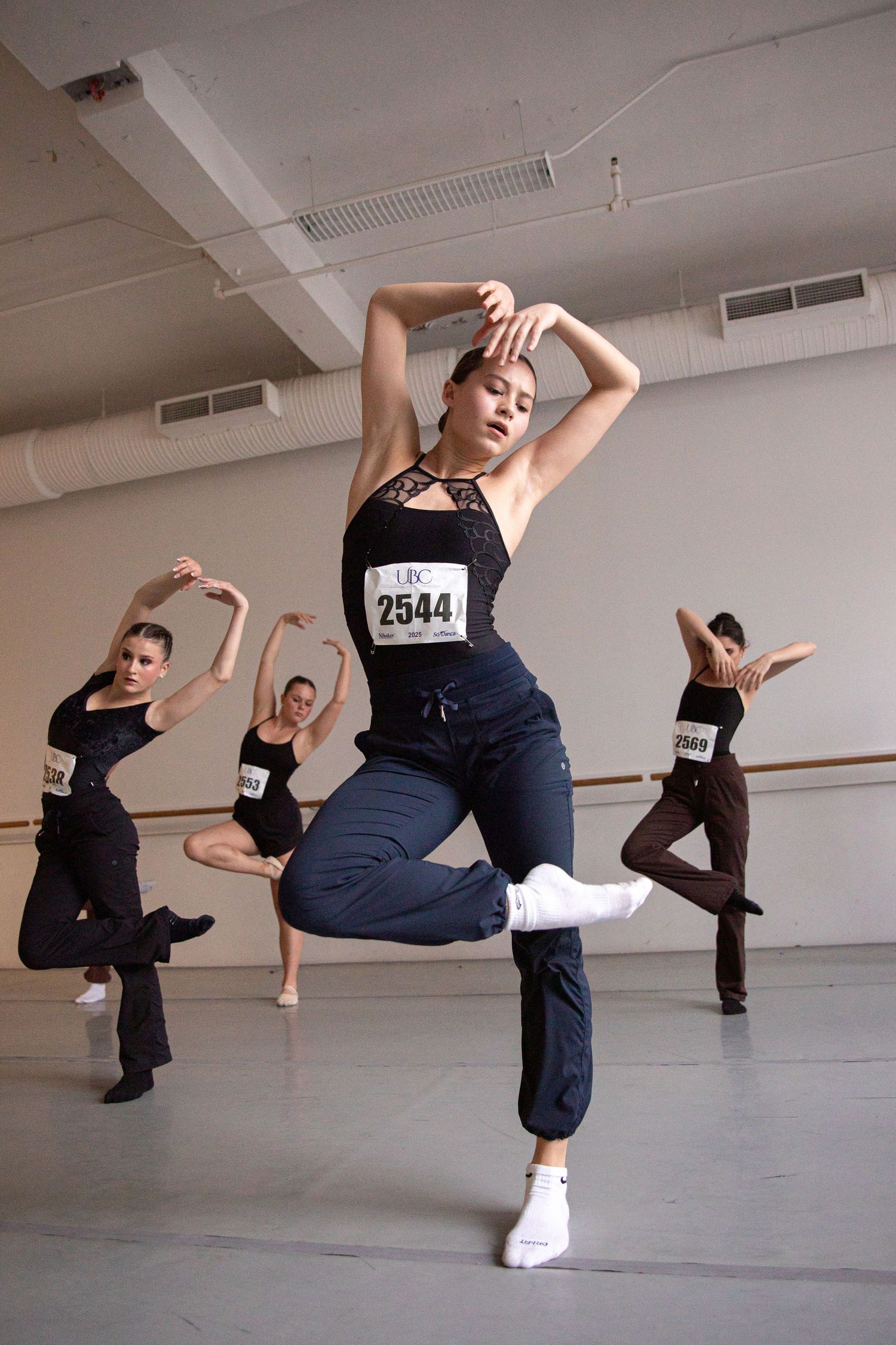 Dancers in numbered bibs practice choreography during dance audition in studio space.