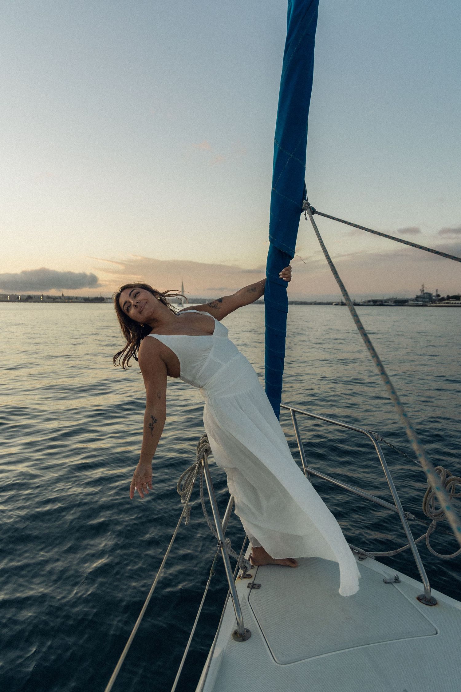 A person in a flowing white dress poses on the deck of a sailboat at sunset over calm ocean waters.