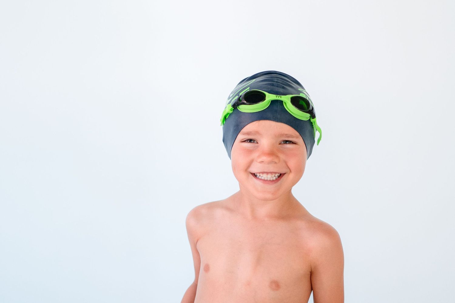 Young swimmer with goggles and swim cap smiling brightly against white background.