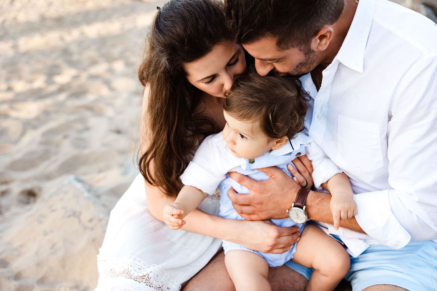 Family of three on the beach, mom and dad kissing their baby boys head