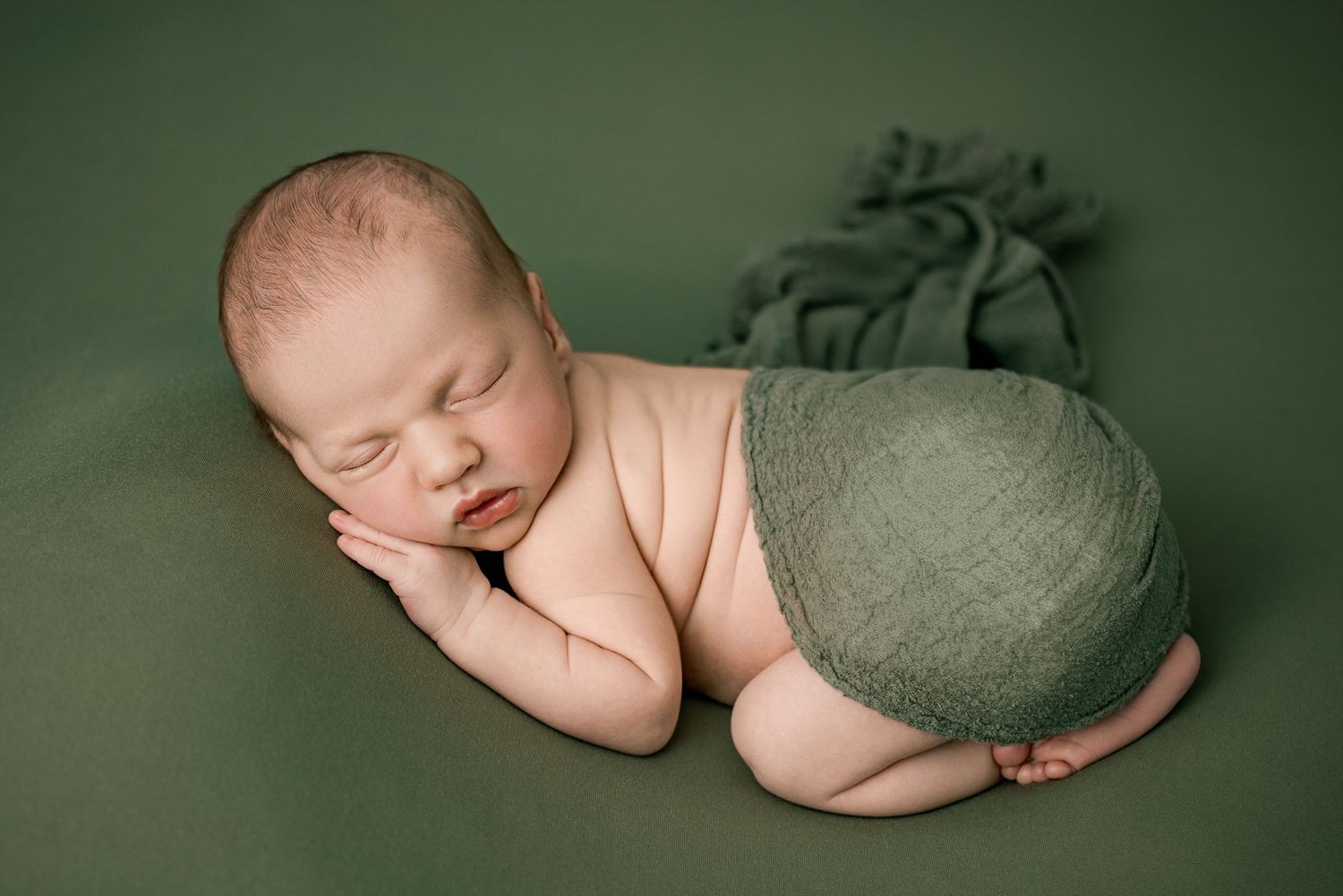 A sleeping newborn wearing soft gray swaddle wrap lies peacefully on green fabric backdrop during professional photoshoot.