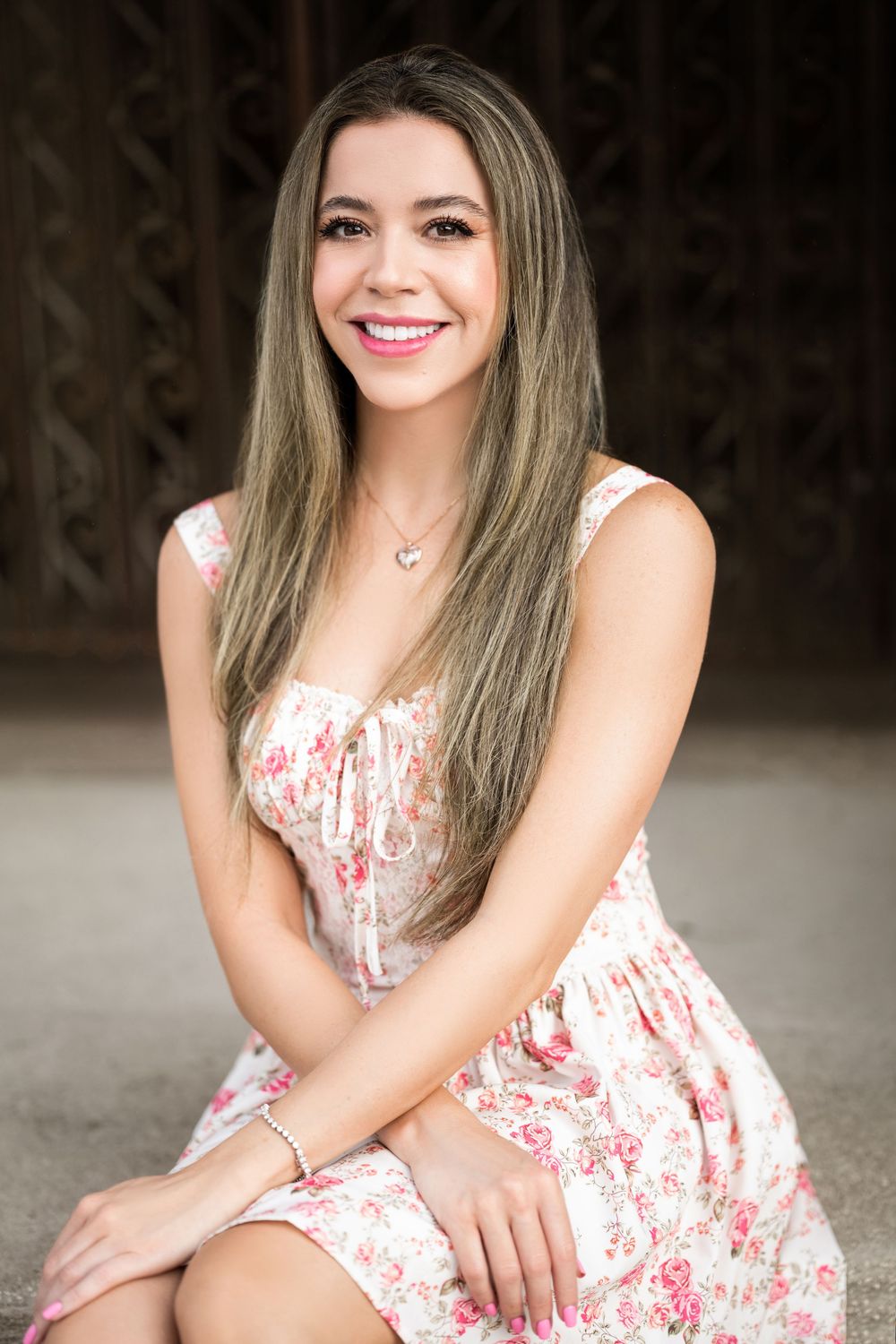 Long-haired woman in floral pink and white summer dress smiling while seated in outdoor setting.