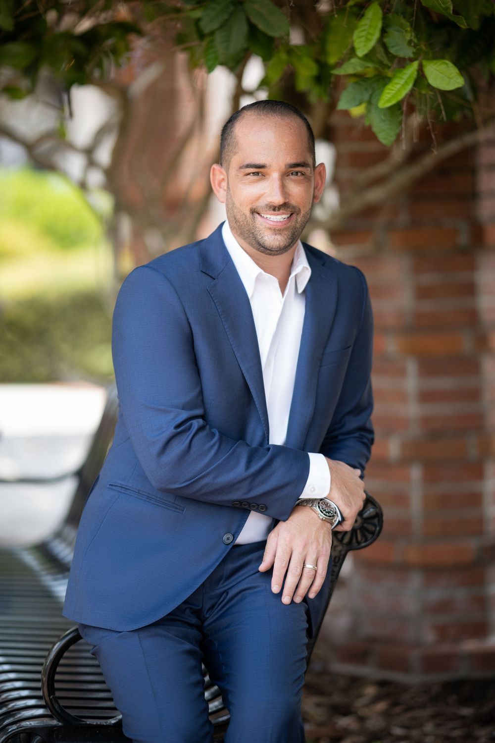 Professional headshot outdoors featuring person in navy suit and white shirt sitting on bench with greenery in background.