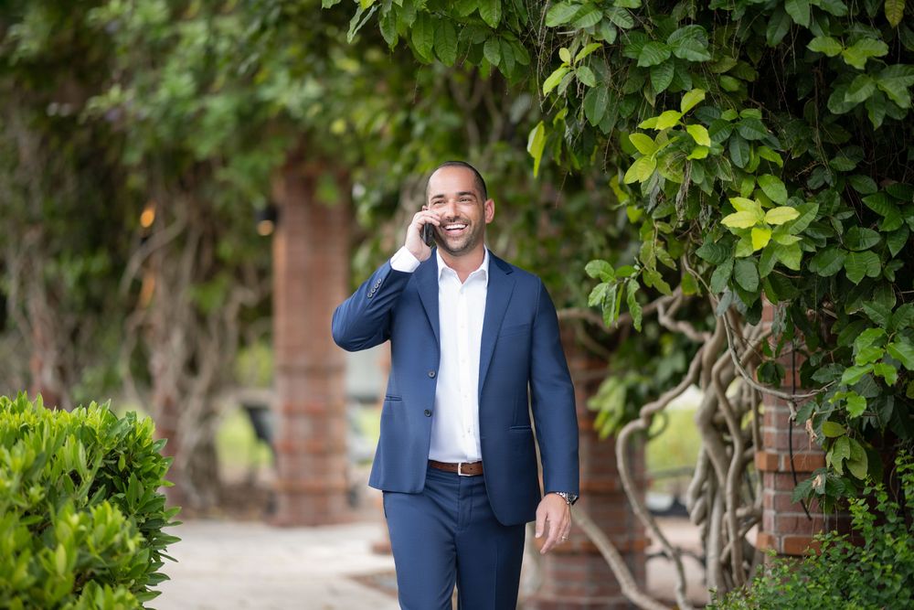 A person in a navy blue suit stands outdoors among lush green foliage and trees in a garden setting.