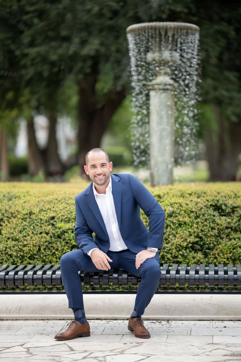 A person in a navy suit sits casually on stone steps with hedges and light decorations in the background.