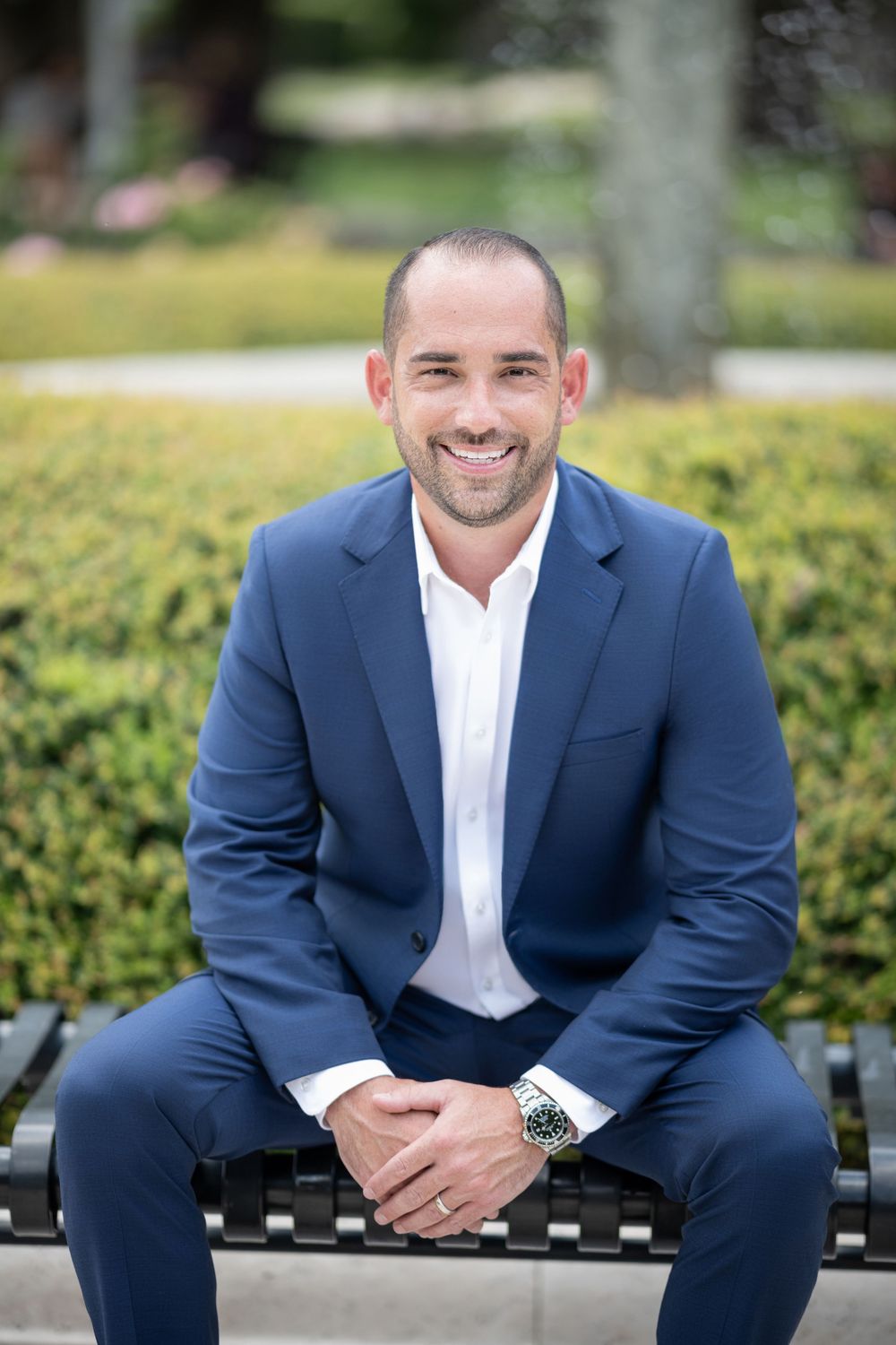 Professional in a navy suit sits on a bench against a lush green hedgerow background.