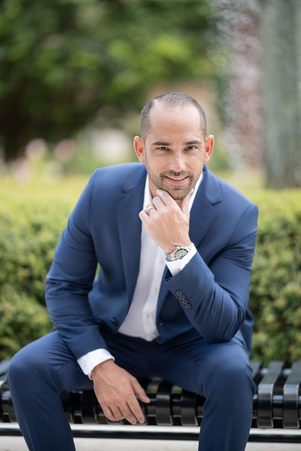 Professional in a navy blue suit sitting on a bench with a thoughtful expression in an outdoor garden setting.