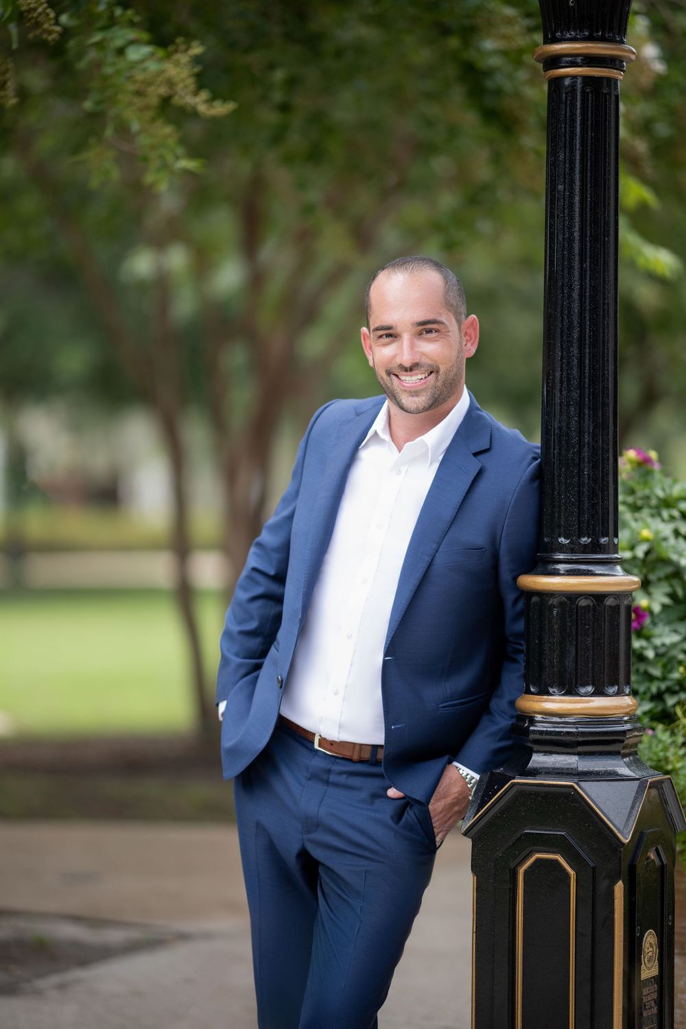 Professional portrait of someone in a navy blue suit and white dress shirt leaning against an ornate lamp post outdoors.