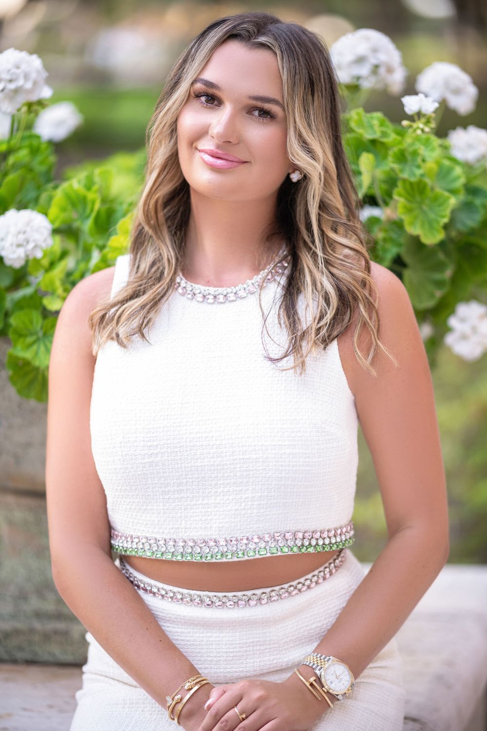A portrait in a white two-piece outfit sitting outdoors with blooming white hydrangeas and greenery in the background.