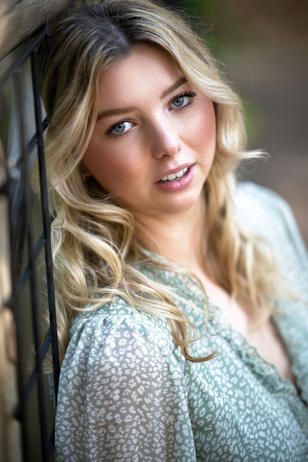 Portrait photograph with soft lighting showing wavy blonde hair, floral print dress, and natural makeup against blurred background.
