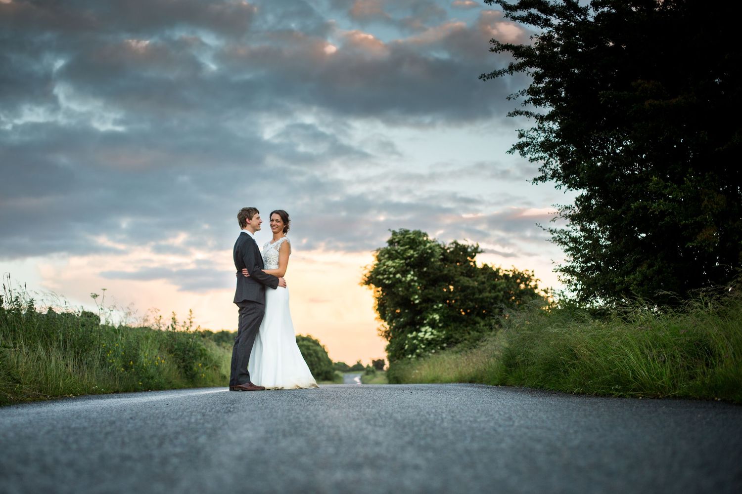 A silhouetted couple stands embracing on a country road at sunset with dramatic cloudy sky.