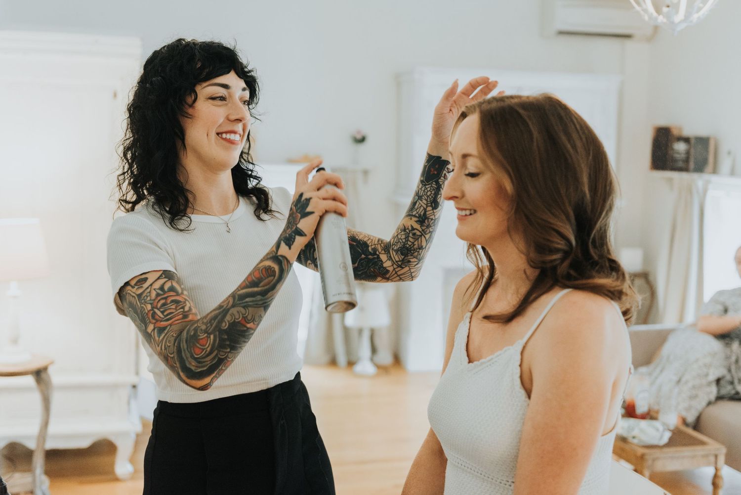 Two laughing friends share a joyful moment in a bright white room, one wearing a sleeve tattoo and white top.