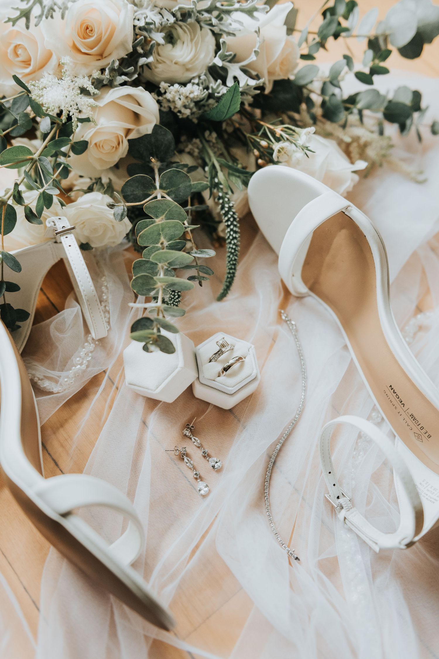 White bridal shoes and delicate cream roses with eucalyptus arranged with hexagonal ring boxes on soft fabric.
