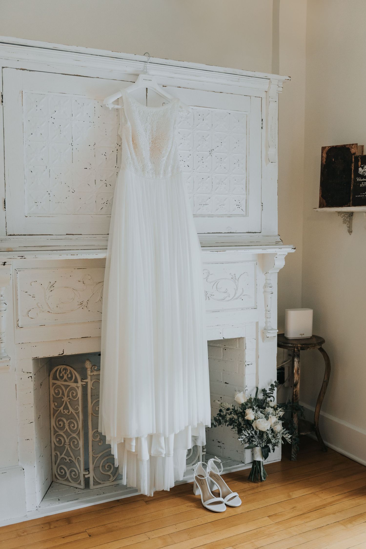 Elegant white wedding dress hangs on vintage fireplace mantle with bridal shoes and floral arrangement below.