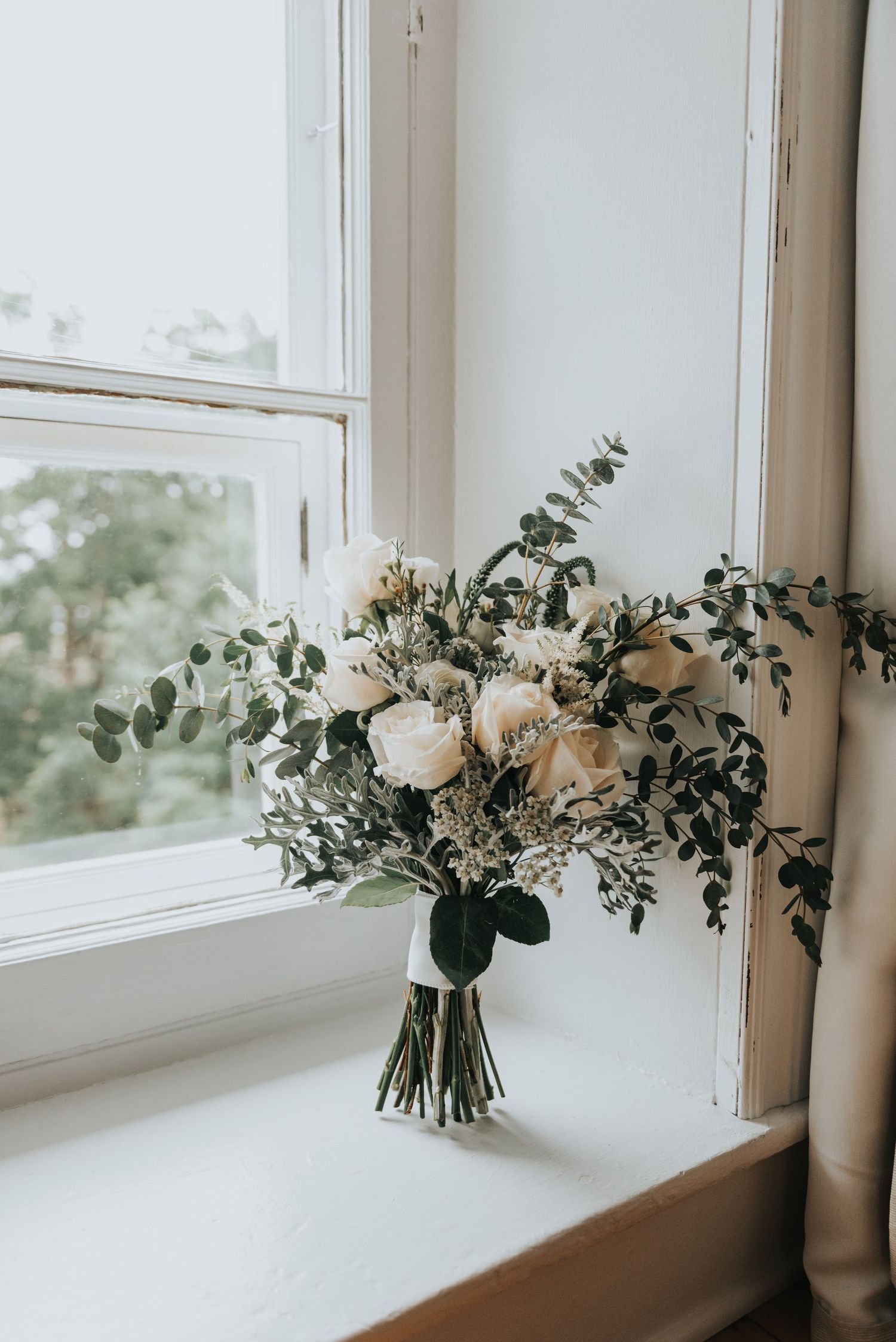 Elegant white rose and eucalyptus bridal bouquet sitting on a windowsill in natural light with moody styling.