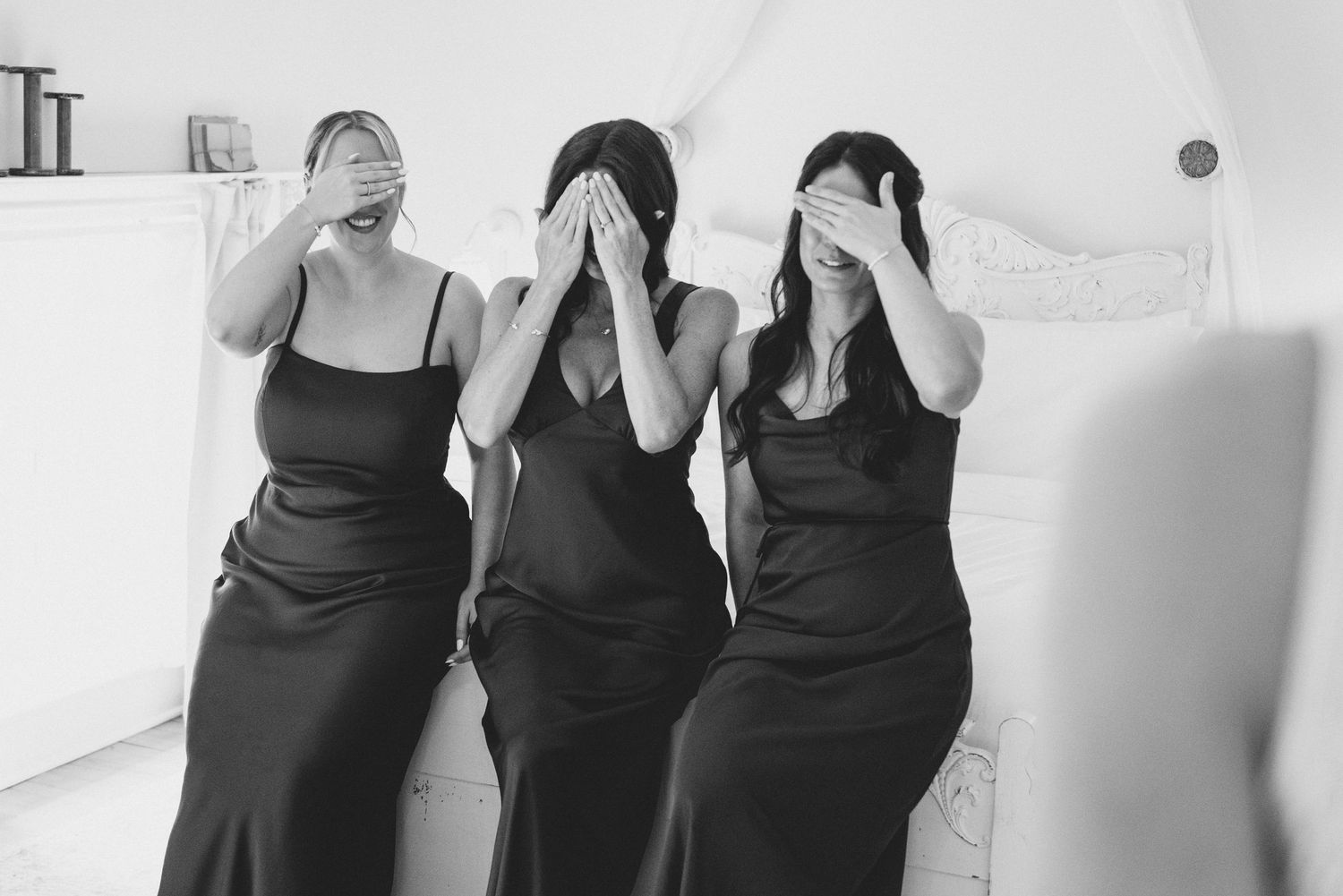 Three women in matching black dresses stand together with their hands covering their eyes in a playful pose.
