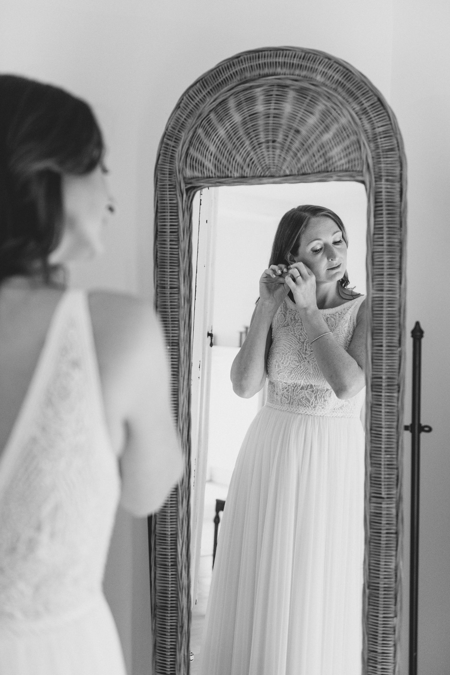 A bride in a flowing white dress puts on earrings while looking in an ornate wicker-framed mirror.