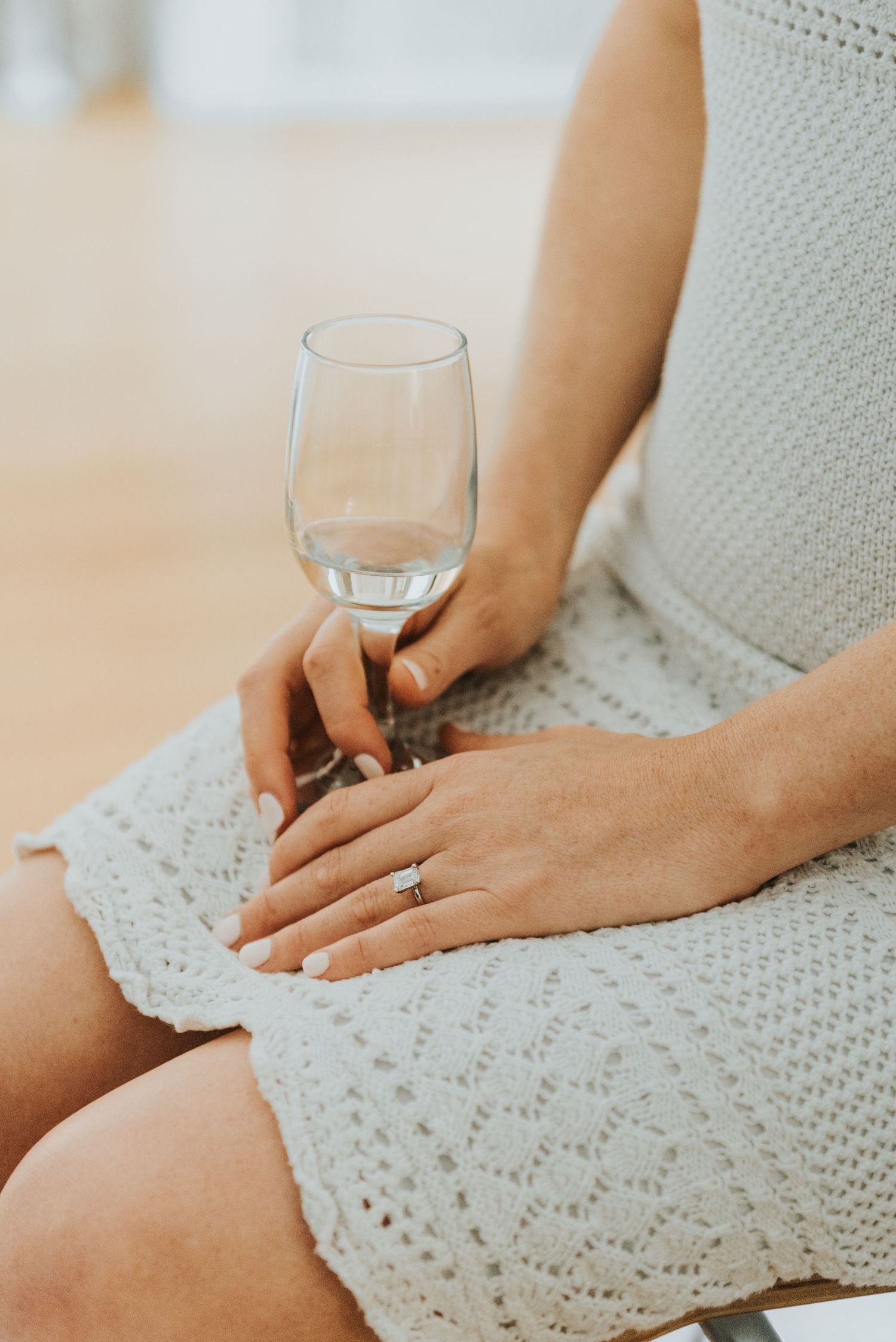 Person in a white lace dress holds a wine glass while sitting, showing an engagement ring on their hand.