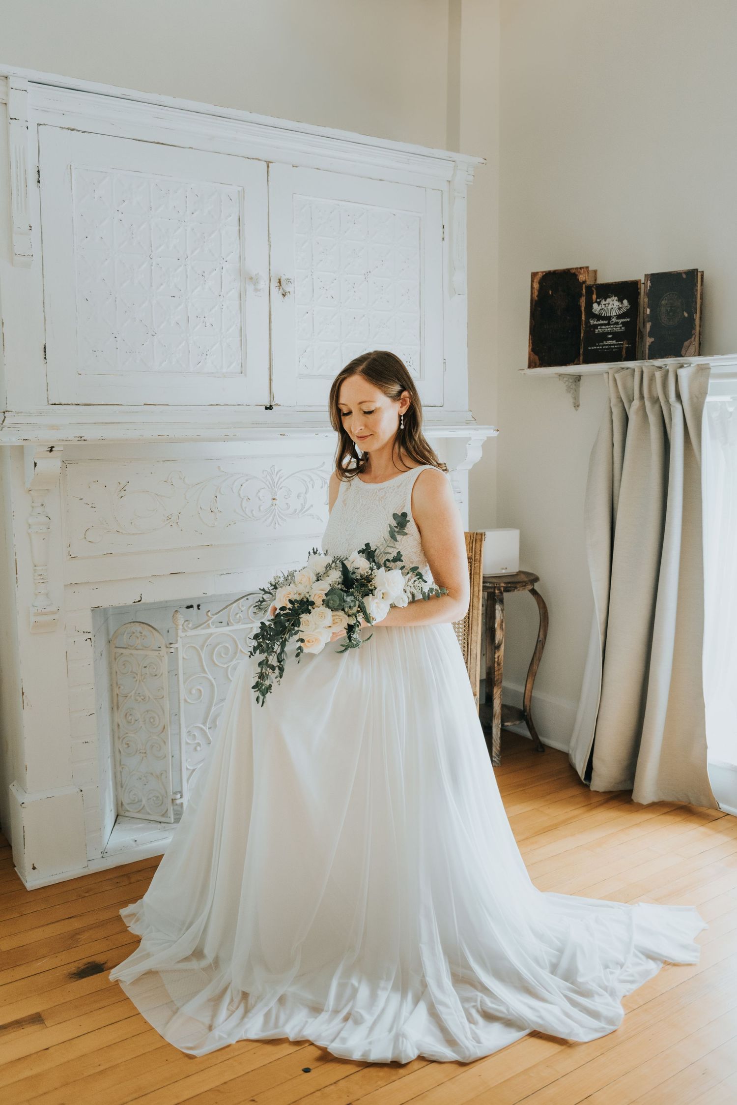 An ornate white wedding dress flows elegantly in a light-filled room with hardwood floors and antique furniture.