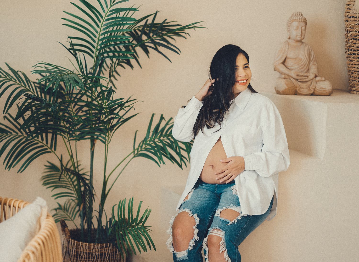 Casual home interior with green palm plant, stone Buddha statue, and relaxed maternity pose against neutral wall.
