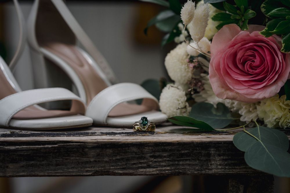 A wedding ring and elegant white sandals rest beside a pink rose bouquet on rustic wooden surface.