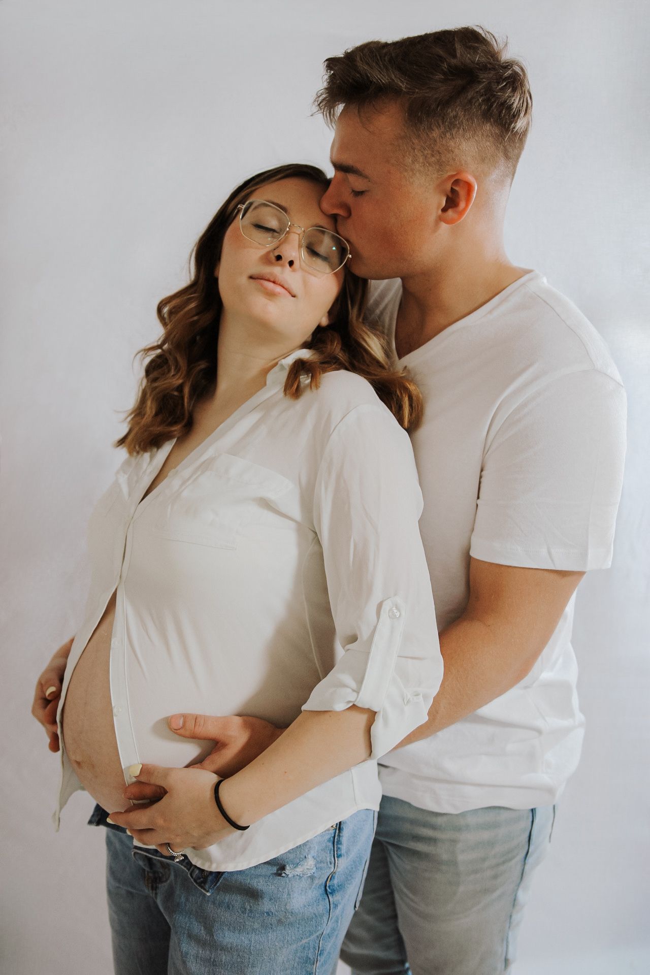 A couple shares tender moments in an intimate white studio setting wearing casual attire.