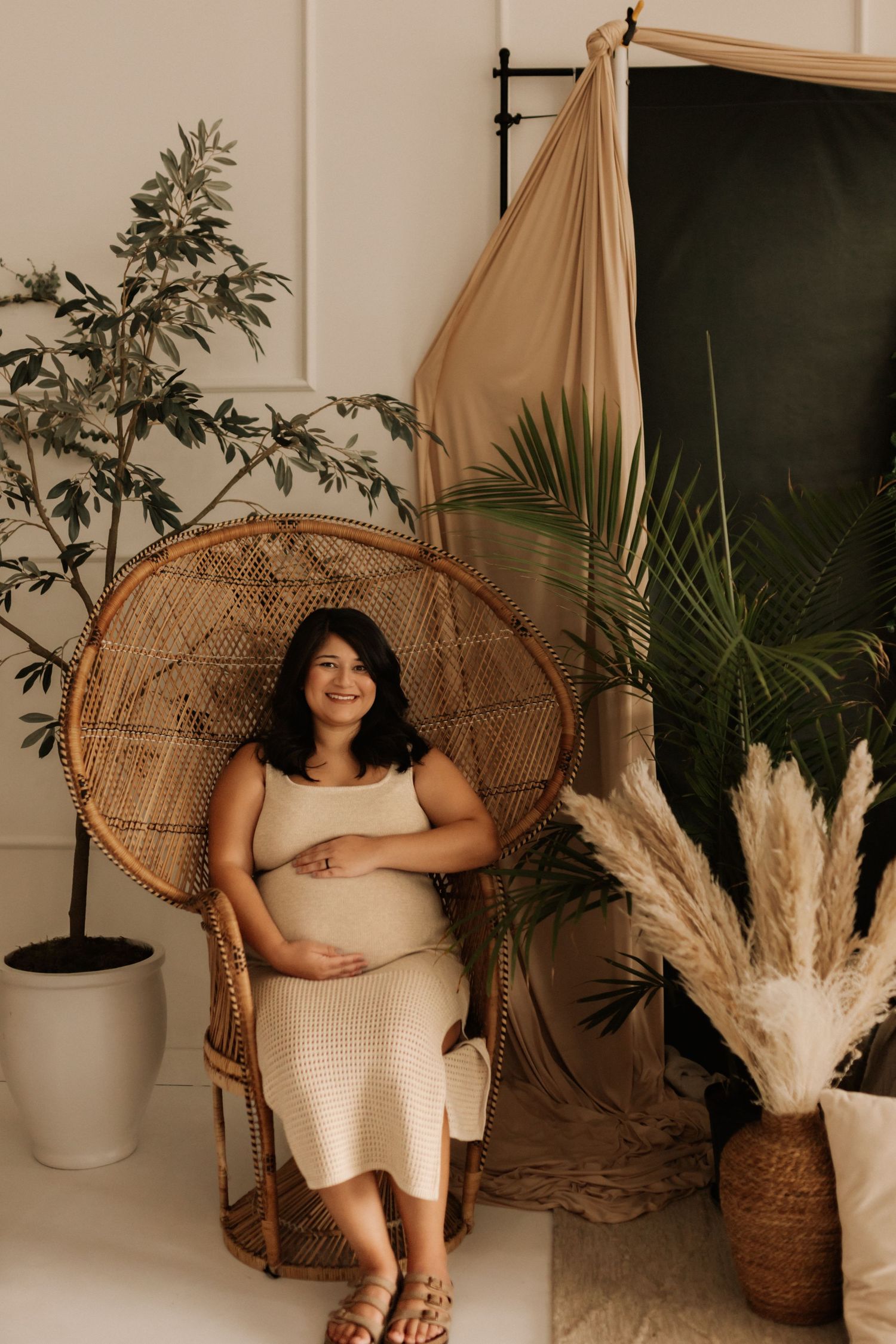 Boho-style indoor maternity photoshoot with peacock chair and tropical plants.