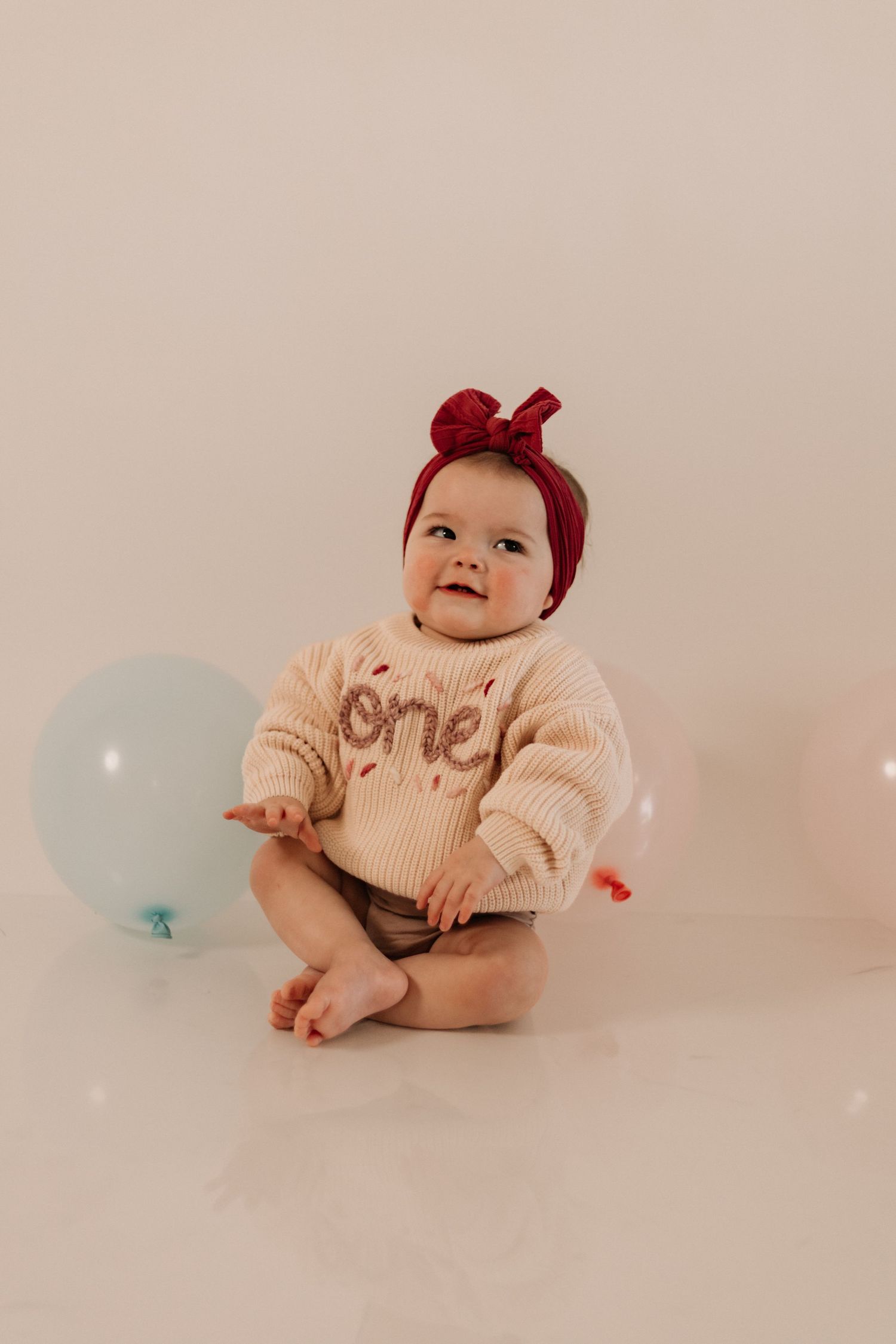 Baby wearing cream sweater and red bow headband posing with blue balloons.