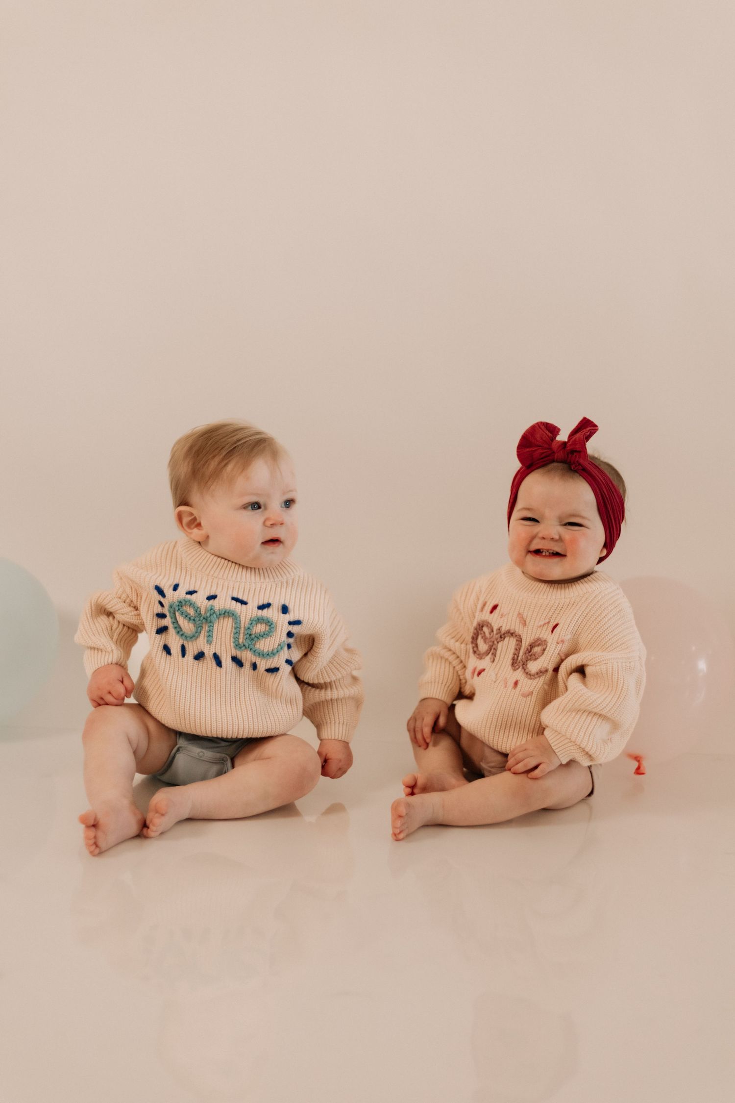 Two babies in matching cream sweaters with one wearing red bow accessory.