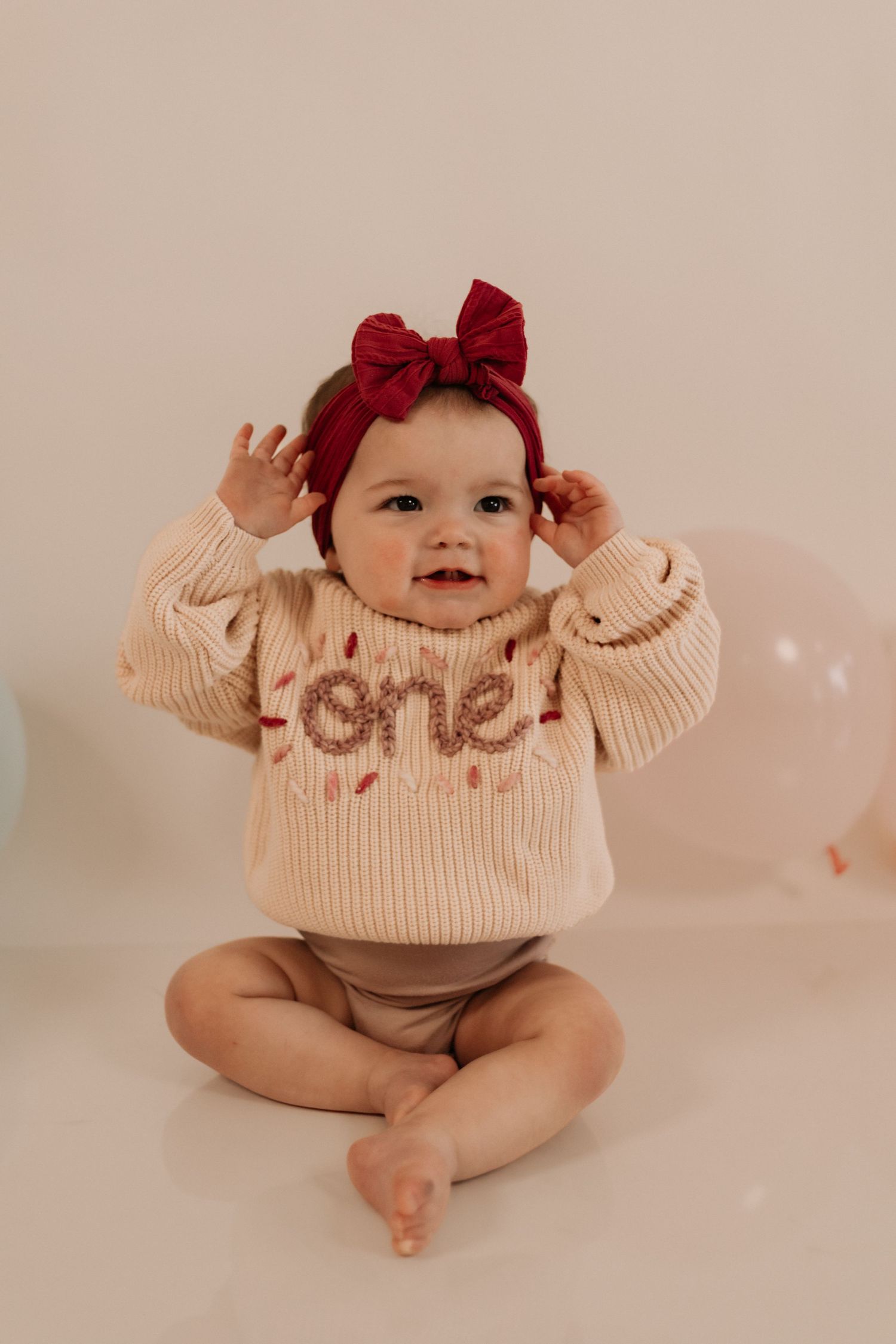 Baby in cream sweater with 'one' text and red bow headband making playful expressions.